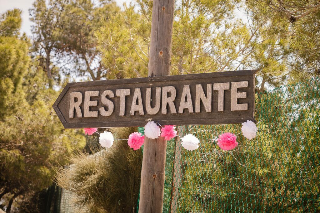 A rustic wooden restaurant sign in Spain, adorned with festive pink and white decorations amidst a natural outdoor setting.